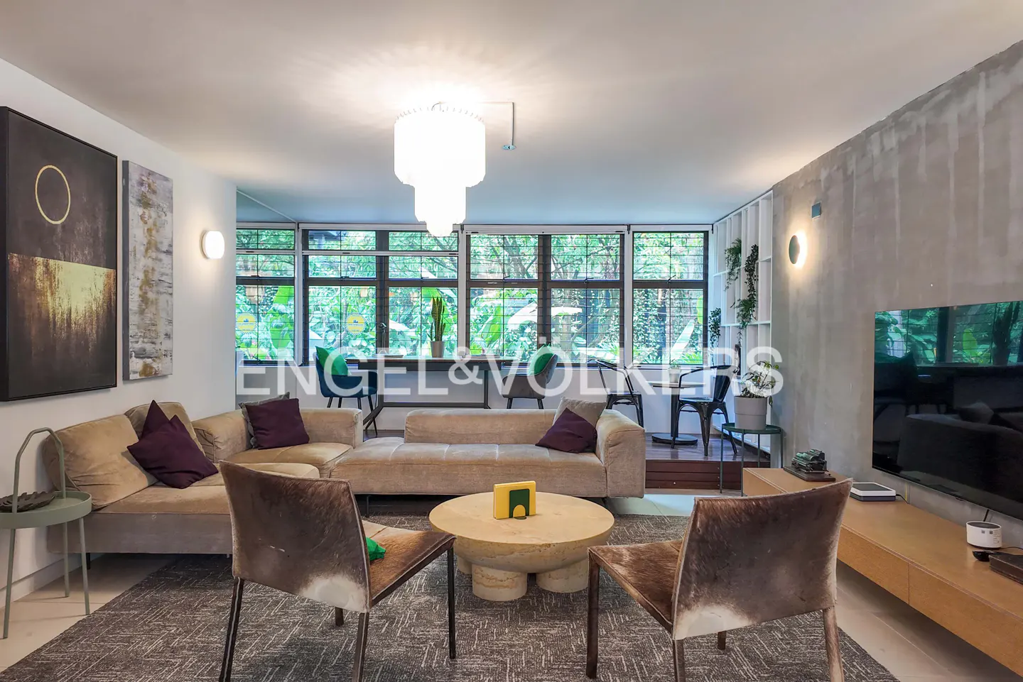 Living room with beige sofas, brown chairs, and a round table on a gray rug. Large windows overlook greenery. Modern chandelier and artwork.