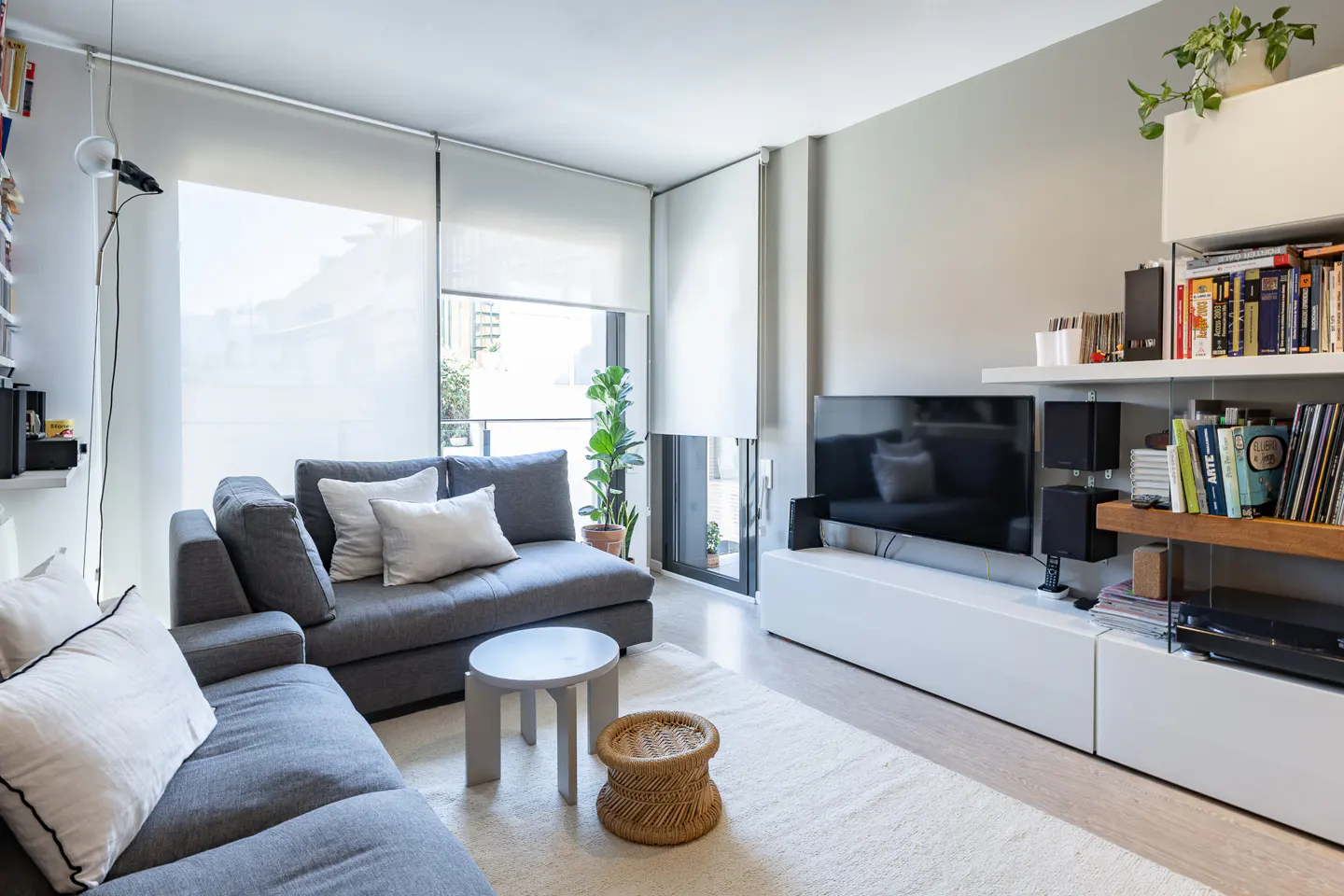 Bright living room with gray sectional sofa, white pillows, round tables, and a white rug. A TV and shelves are on the right.