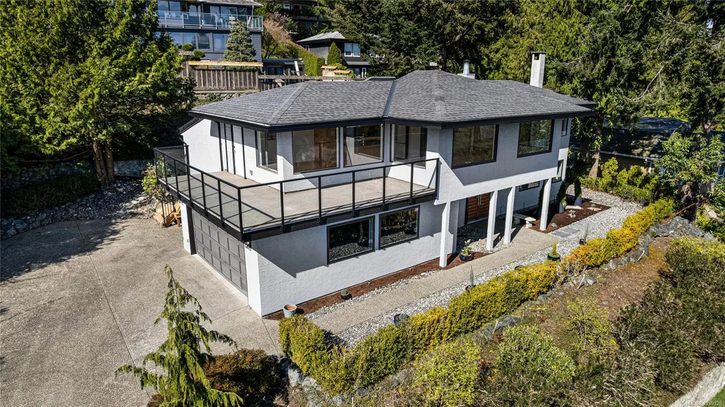 Two-story white house with a gray roof, black trim, and a balcony, surrounded by green trees and bushes.