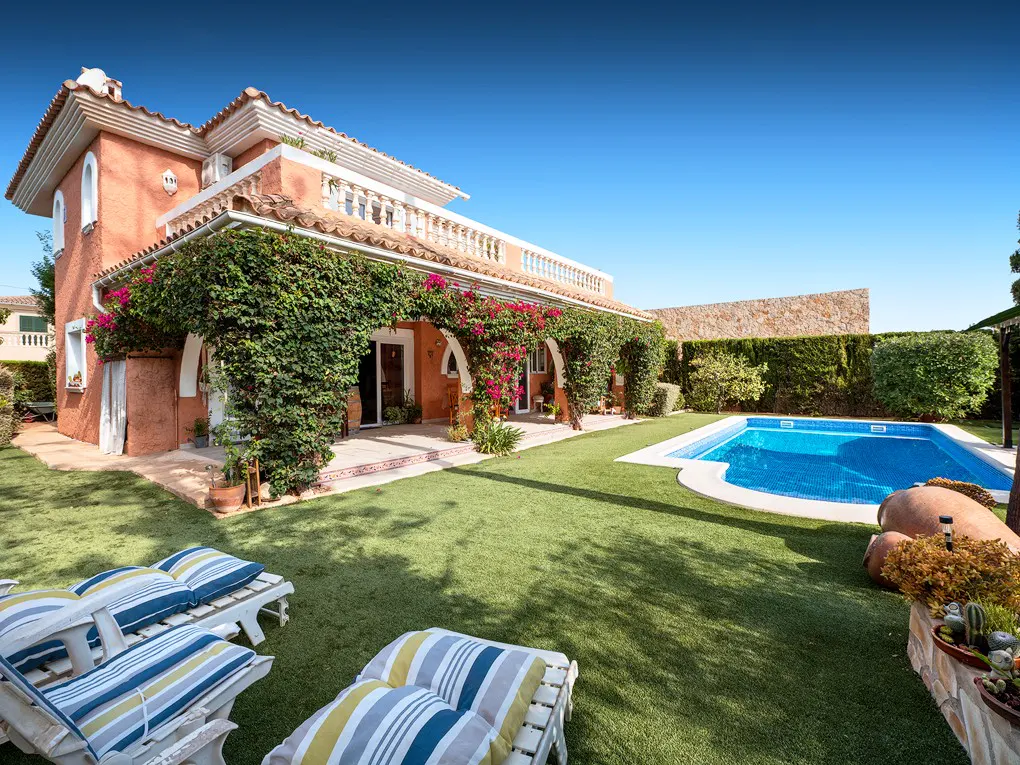Exterior of a peach-colored house with a pool, green lawn, and lounge chairs under a clear blue sky.