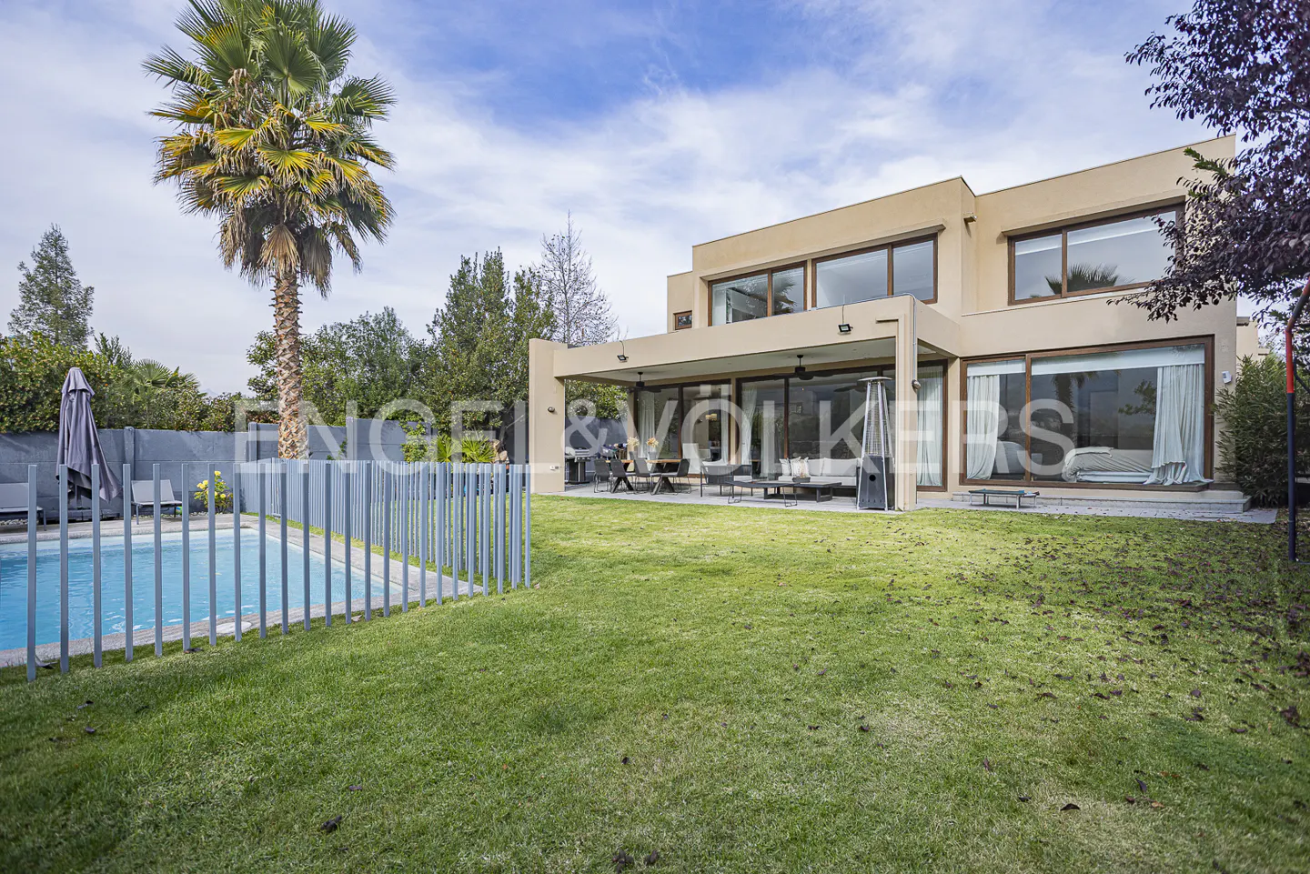 Tan two-story house with large windows, a patio, and a pool surrounded by a gray fence. A palm tree stands to the left.