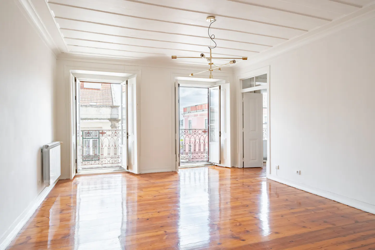 Bright, empty room with wood floors, white walls, and two open balcony doors. A modern chandelier hangs from the white, paneled ceiling.