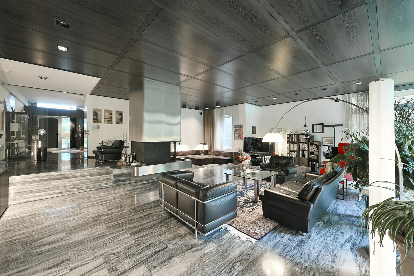 A modern living room with black leather sofas, a metal fireplace, and a dark wood-paneled ceiling.