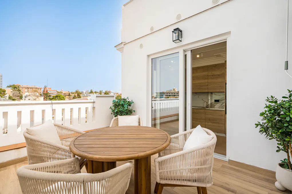 A bright balcony with a round wooden table and four woven chairs with white cushions. Sliding glass doors lead to a kitchen. City view in the background.