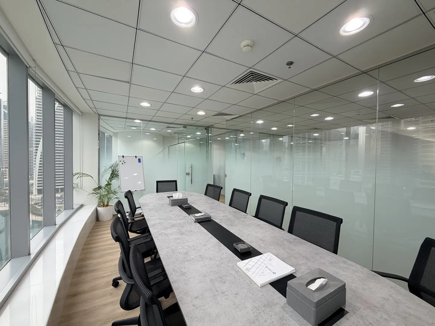 Bright, modern office conference room with a long gray table, black chairs, and frosted glass walls. A whiteboard and plant are near a large window.