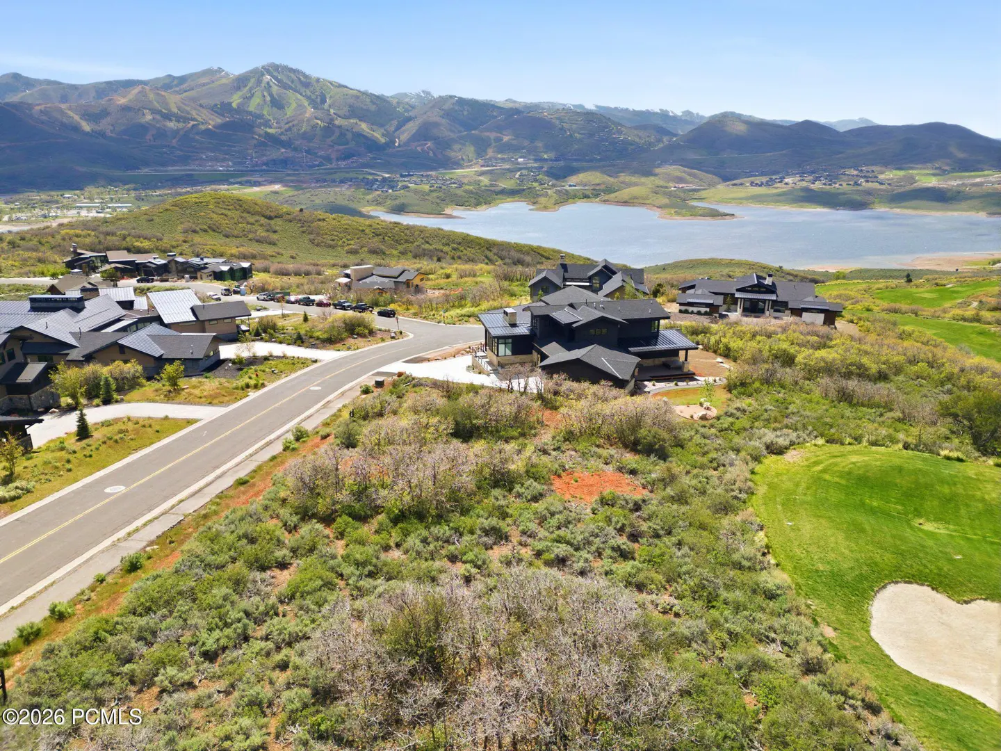 Aerial view of modern homes with dark roofs, a road, and a golf course, set against a backdrop of mountains and a lake.
