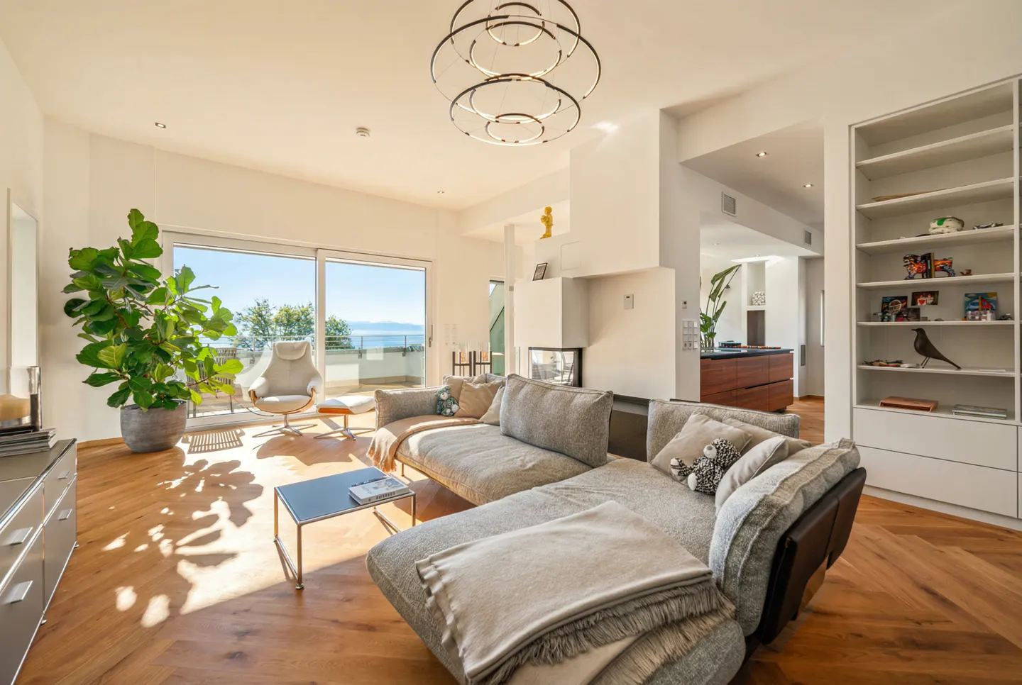 Bright living room with a gray sectional sofa, wood floors, and a large window with a view of the ocean. A modern chandelier hangs from the ceiling.