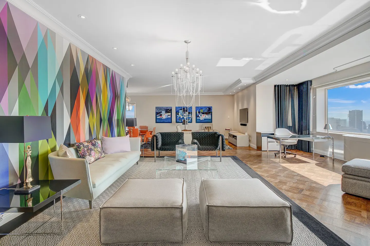 Bright living room with colorful geometric wall art, light sofa, and two gray ottomans. A chandelier hangs above a glass coffee table and two black chairs. A desk sits by a window with a city view.