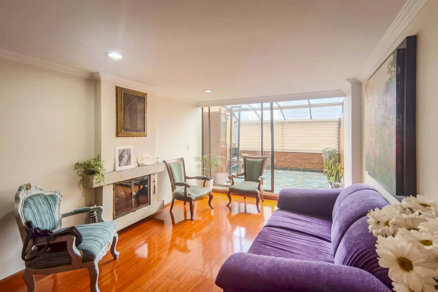 Bright living room with hardwood floors, purple sofa, and green chairs. A fireplace and art adorn the walls, leading to a sunlit patio.