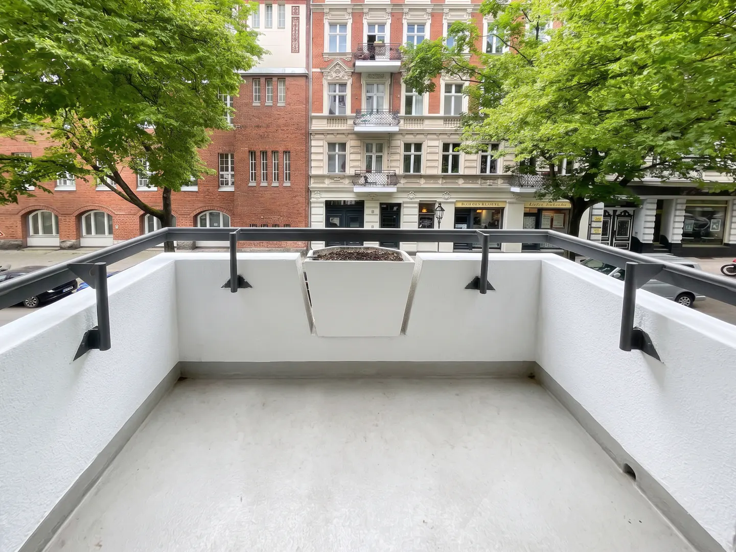 A white balcony with a black railing overlooks a city street with brick buildings and green trees. A white planter box is attached to the balcony wall.