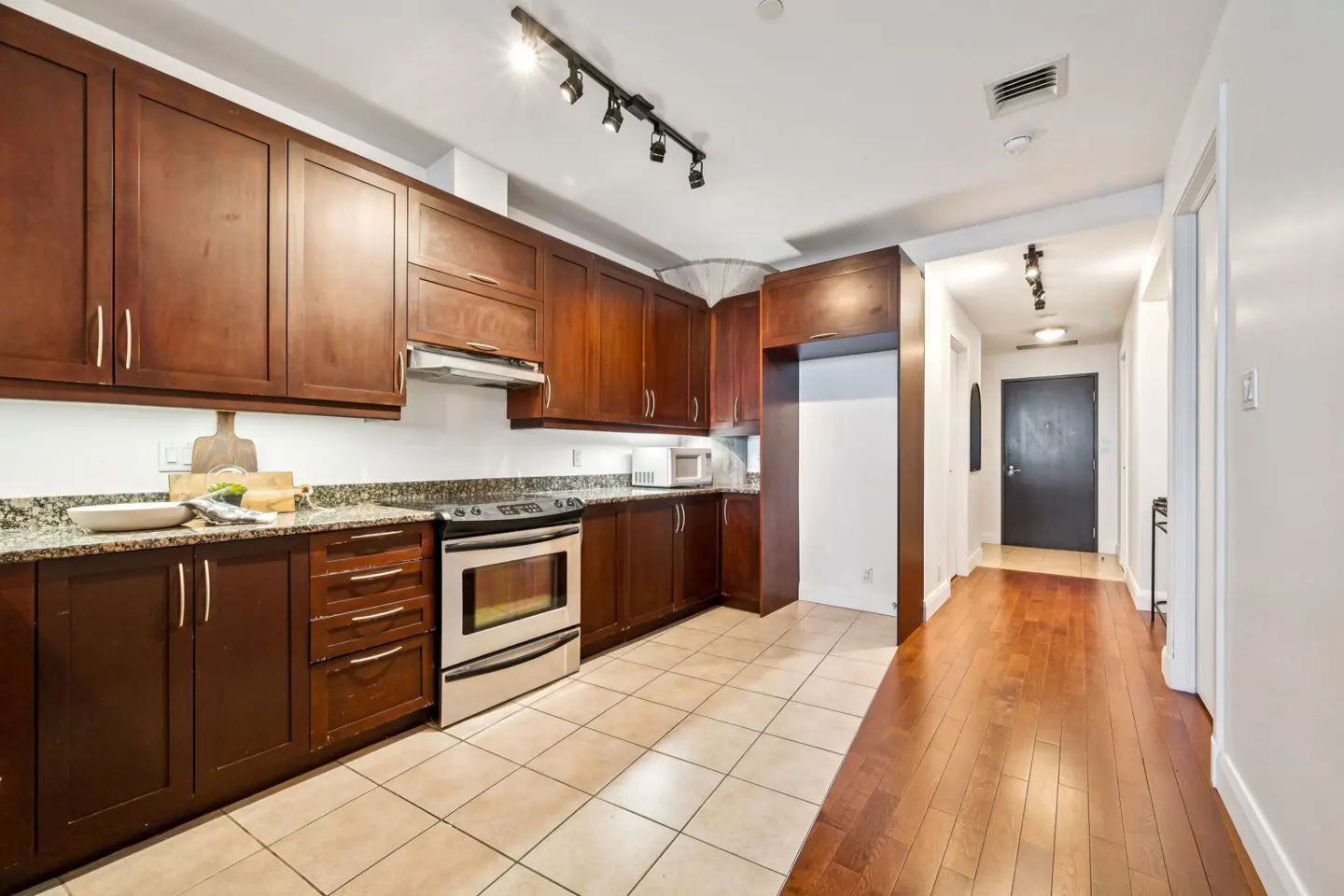 A kitchen with dark wood cabinets, stainless steel appliances, and granite countertops. The floor is tiled, transitioning to wood in the hallway.