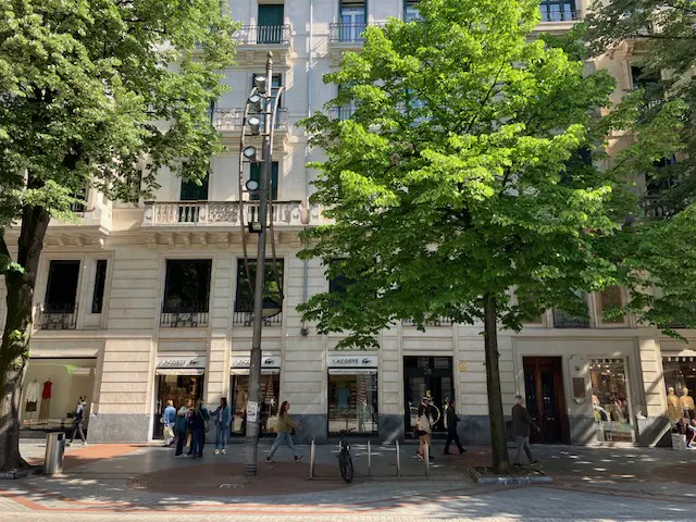 Street view of a Lacoste store in a light stone building with green trees and people walking by.
