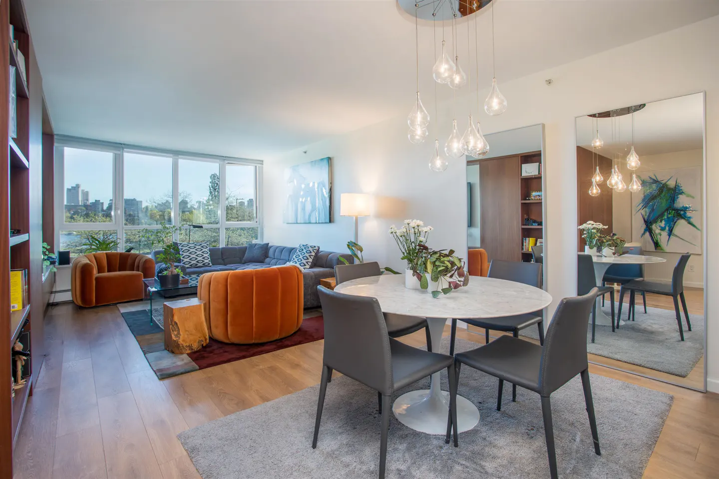 Bright, modern living and dining area with orange chairs, gray sofa, and round white table under a glass pendant light fixture.