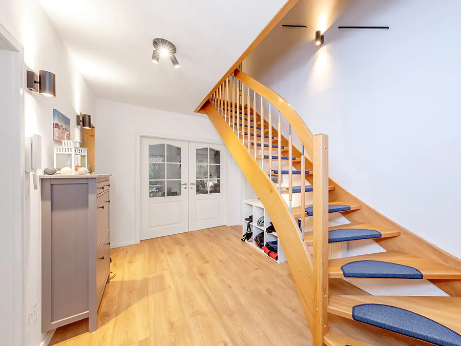 Bright foyer with wood floors, white walls, and a light wood staircase with blue treads. A gray cabinet sits near white double doors.