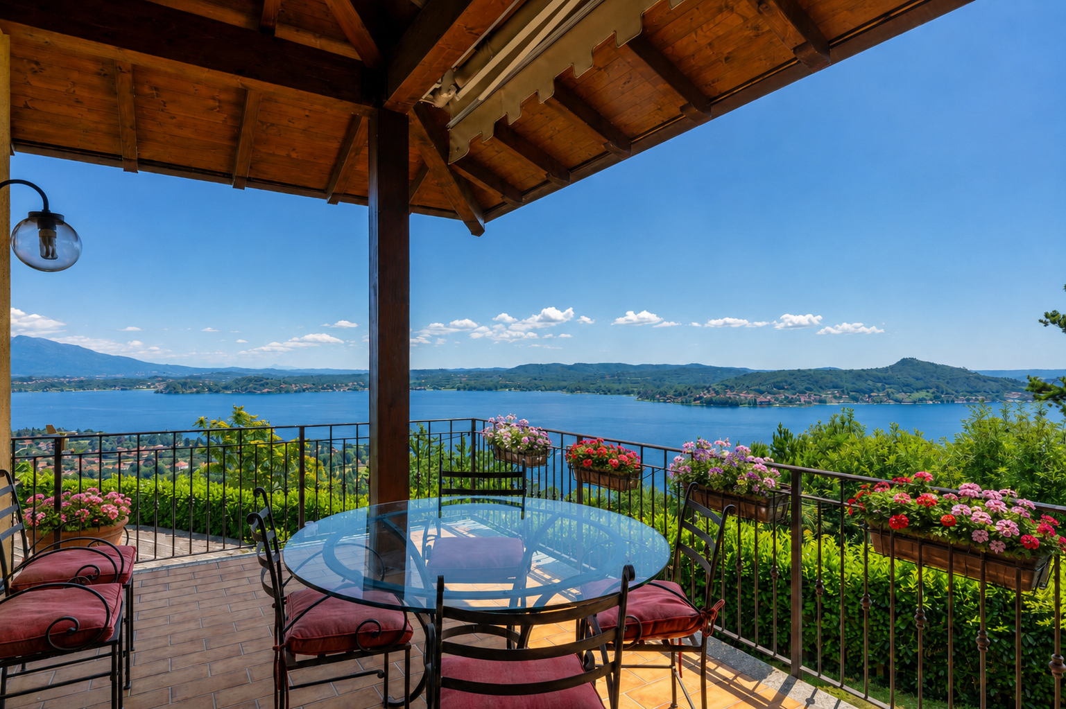 Covered terrace with a glass table, chairs, and flower boxes overlooking a scenic lake and mountains under a blue sky.