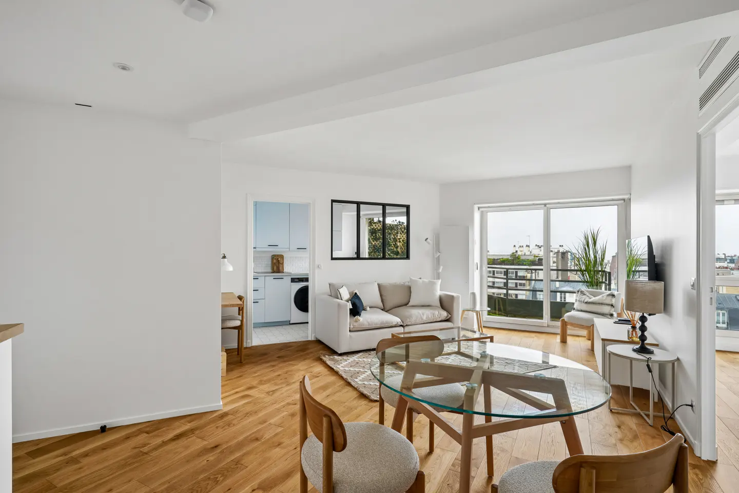 Bright, modern apartment interior with hardwood floors, white walls, and a glass-top dining table. A beige sofa faces a balcony with city views.