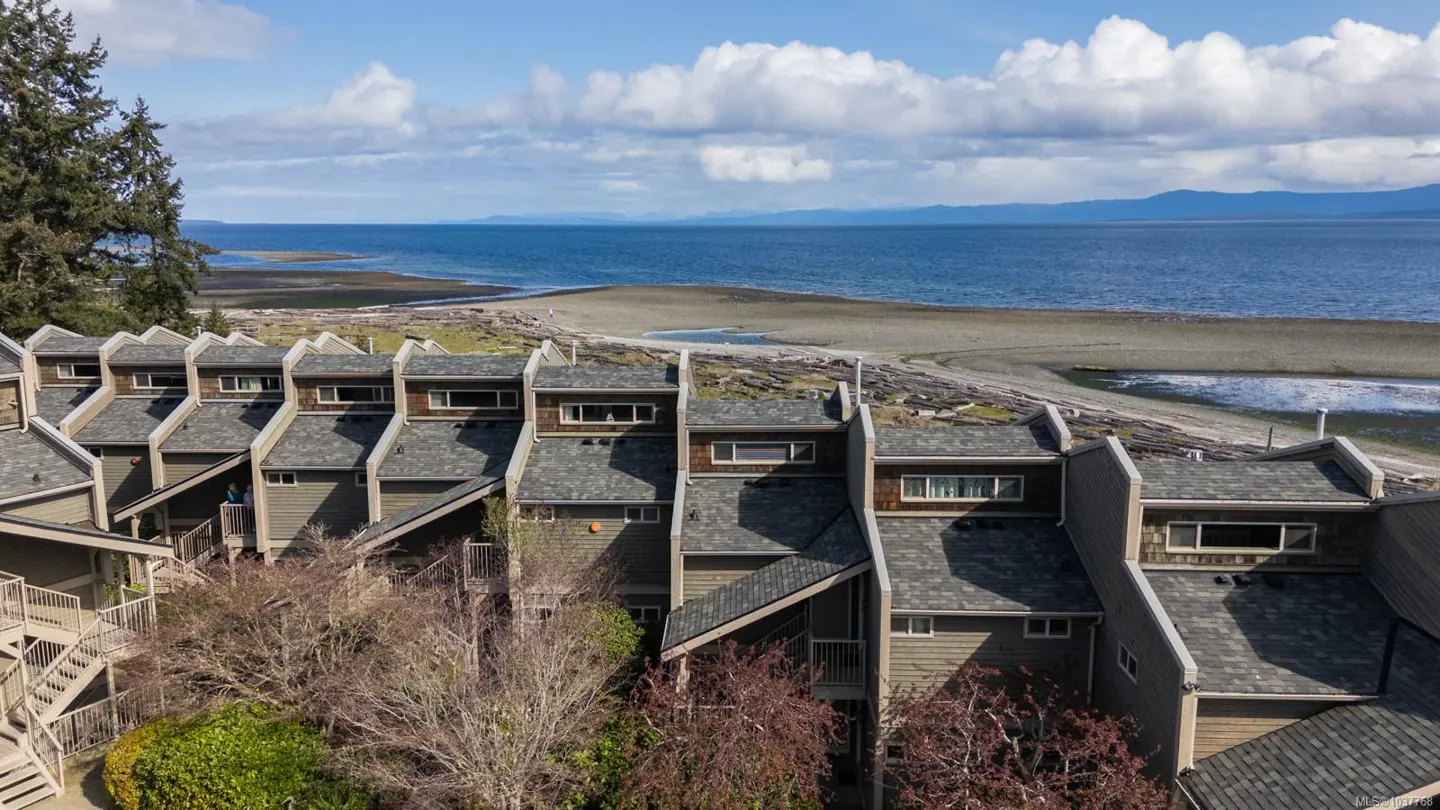 Aerial view of beachfront townhouses with gray roofs, tan siding, and ocean backdrop under a blue sky with scattered clouds.