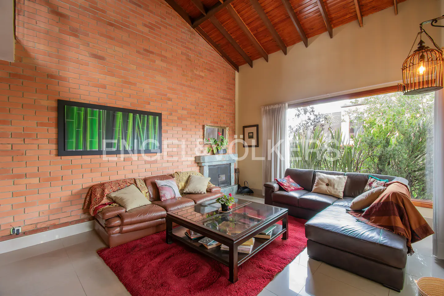 Living room with brick wall, brown leather sofas, and a red rug. A fireplace and large window with greenery outside.