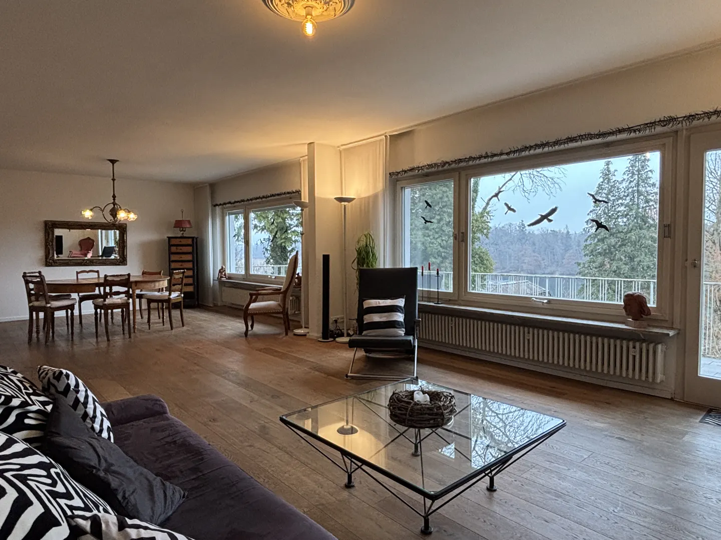 A spacious living room with hardwood floors, a dining table, a sofa, and large windows overlooking a forest.