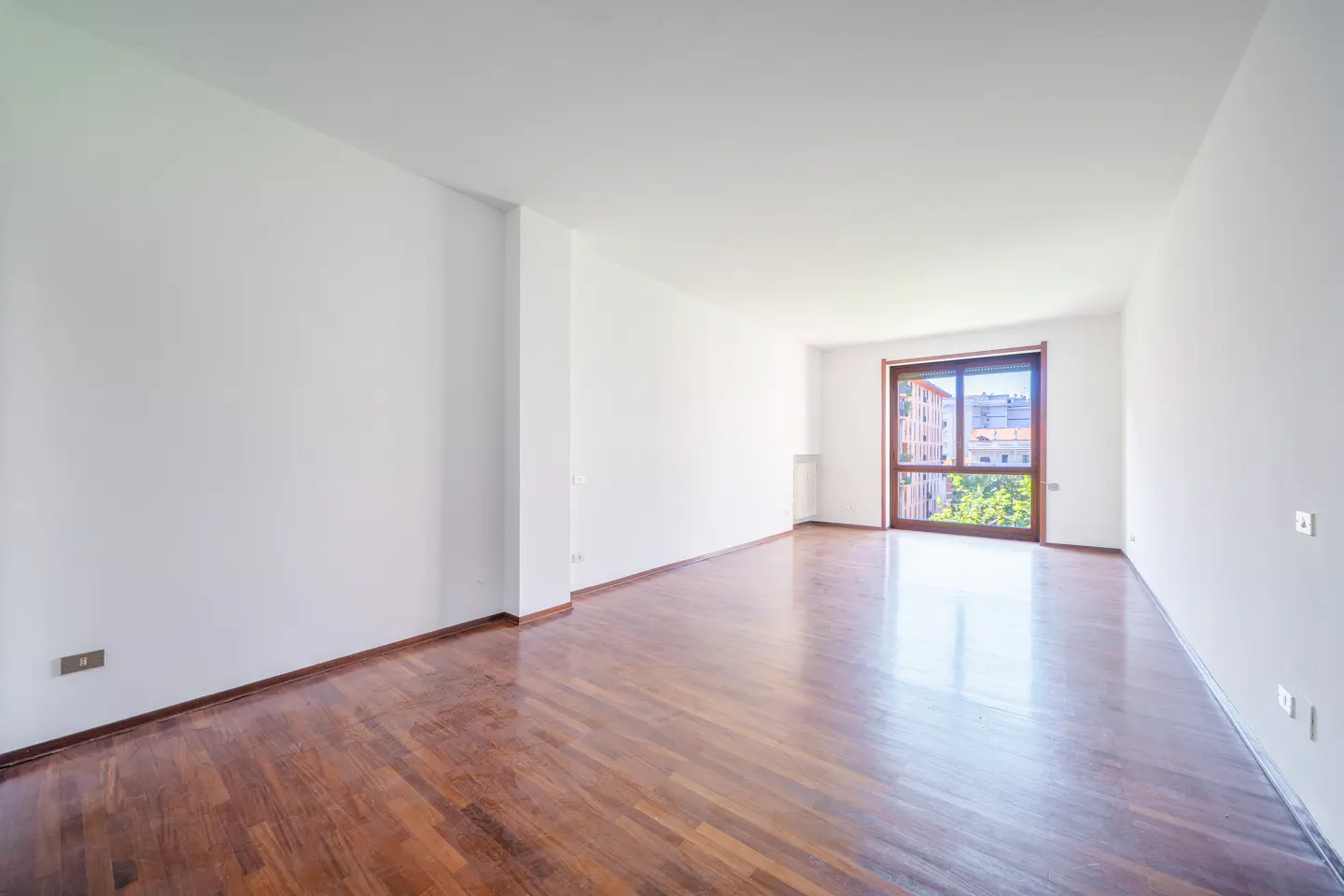 Bright, empty room with white walls and a dark wood floor. A large window shows a view of a building and trees.