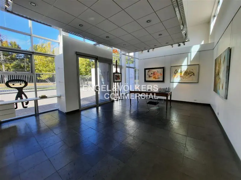 Bright gallery space with large windows, dark tile floor, and white walls. Artwork hangs on the walls, and a table and chair sit in the center.