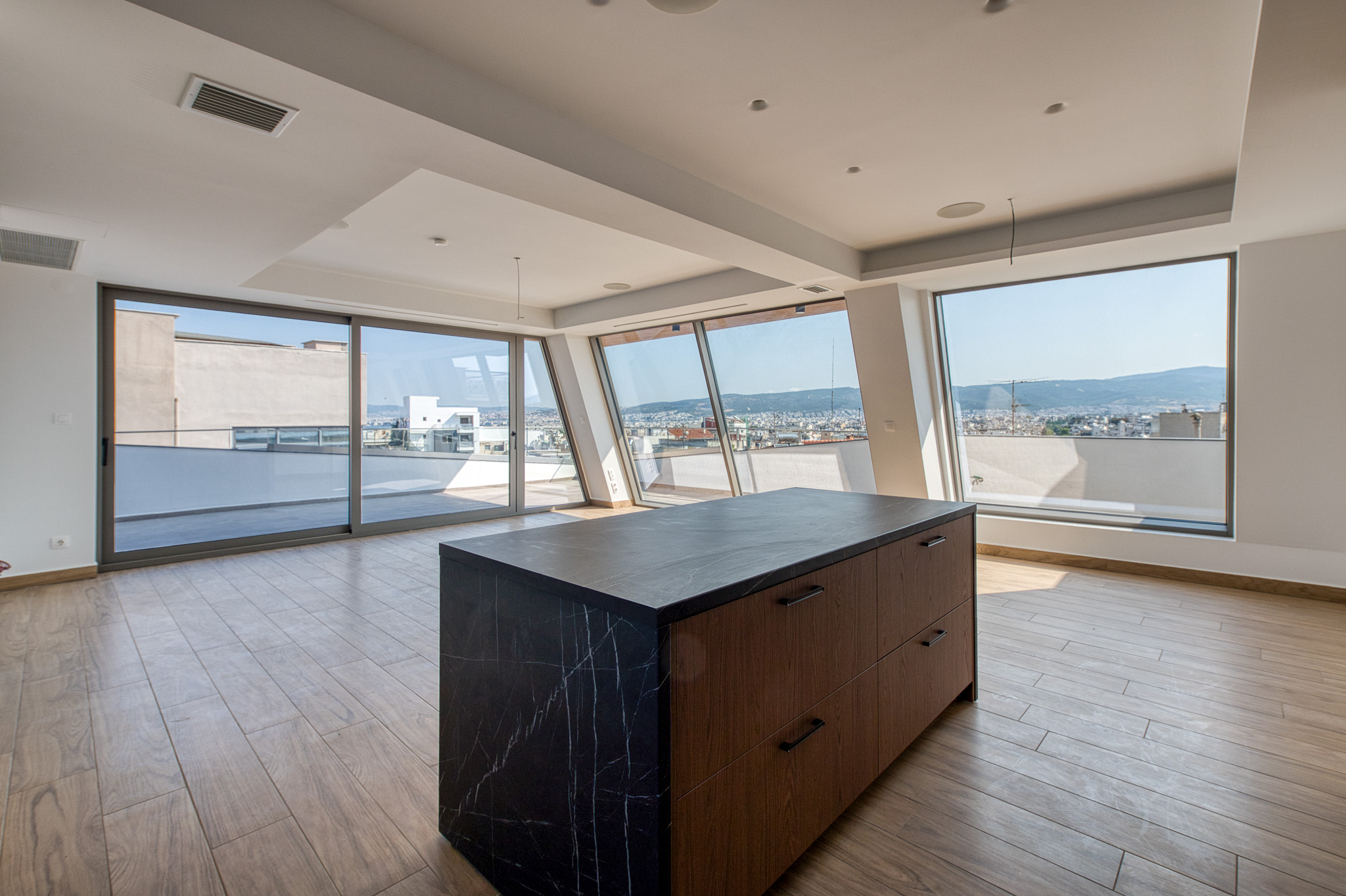 Modern kitchen island with black marble and wood cabinets in a bright room with floor-to-ceiling windows and city views.