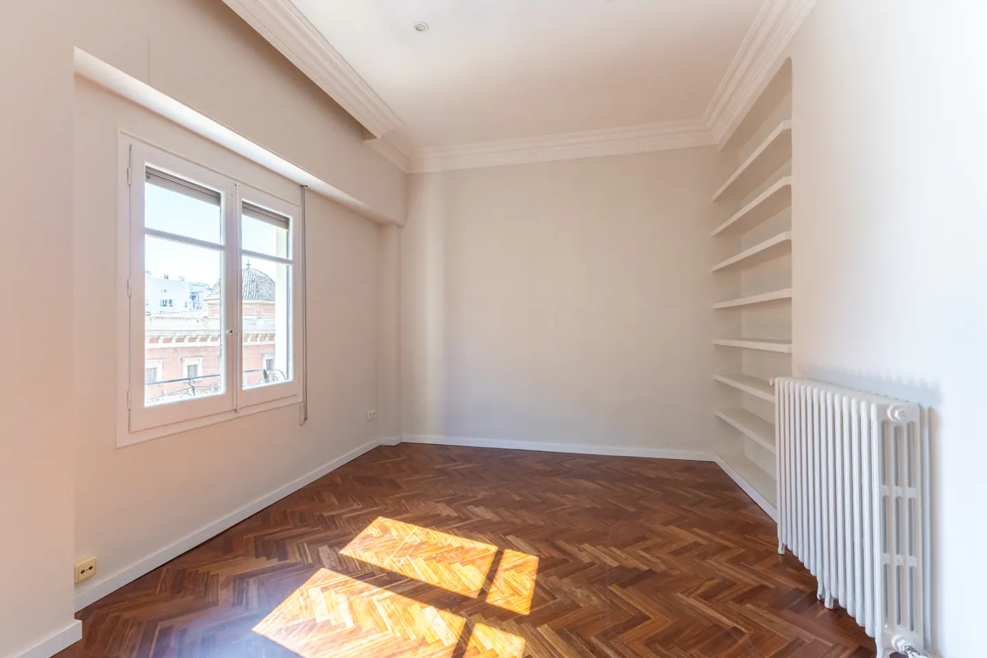 Bright, empty room with herringbone wood floors, white walls, and a window. Built-in shelves and a radiator are on the right wall.