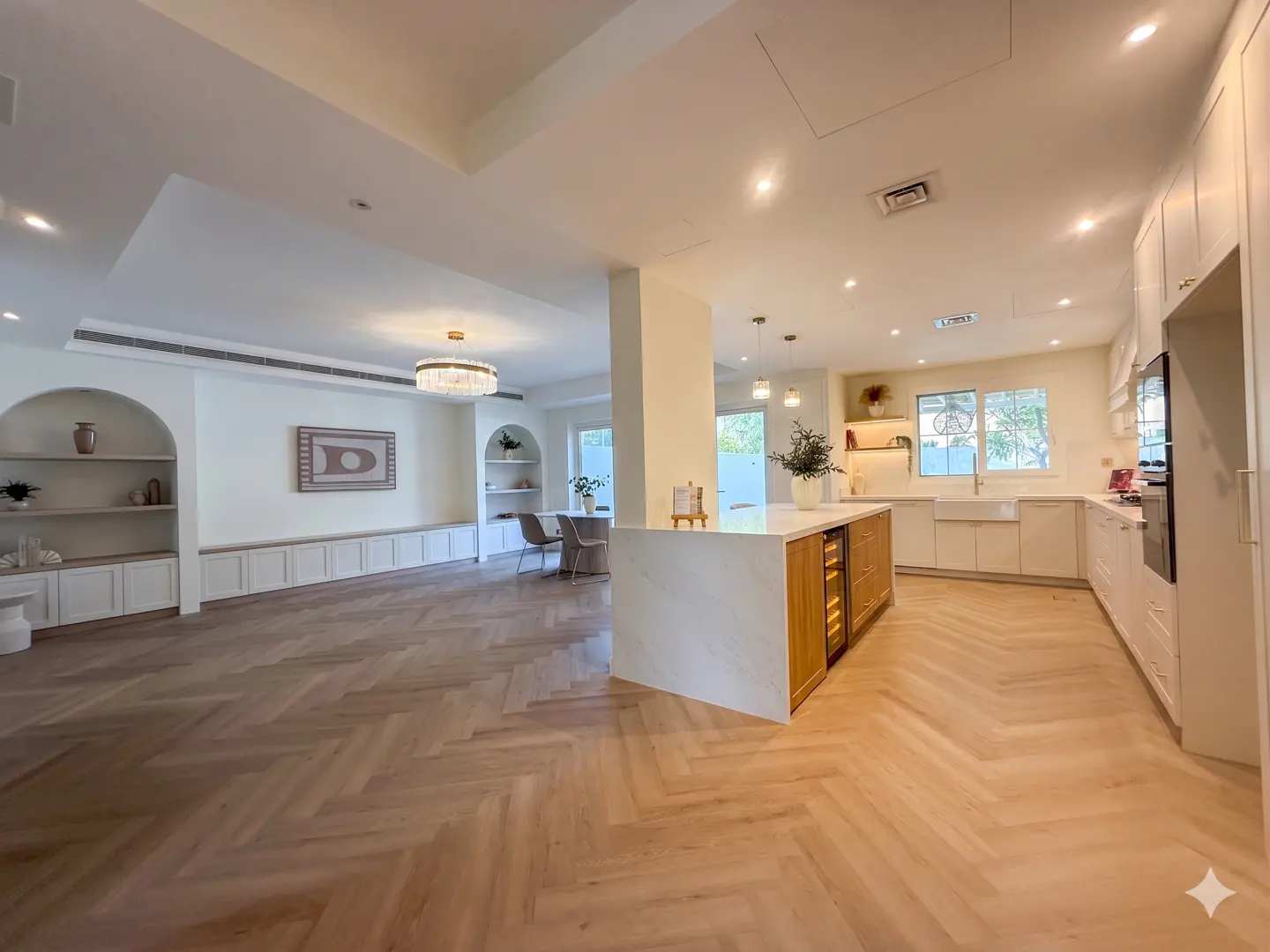 Bright, open-concept kitchen and living area with herringbone wood floors. White cabinets, island with wine fridge, and built-in shelving.