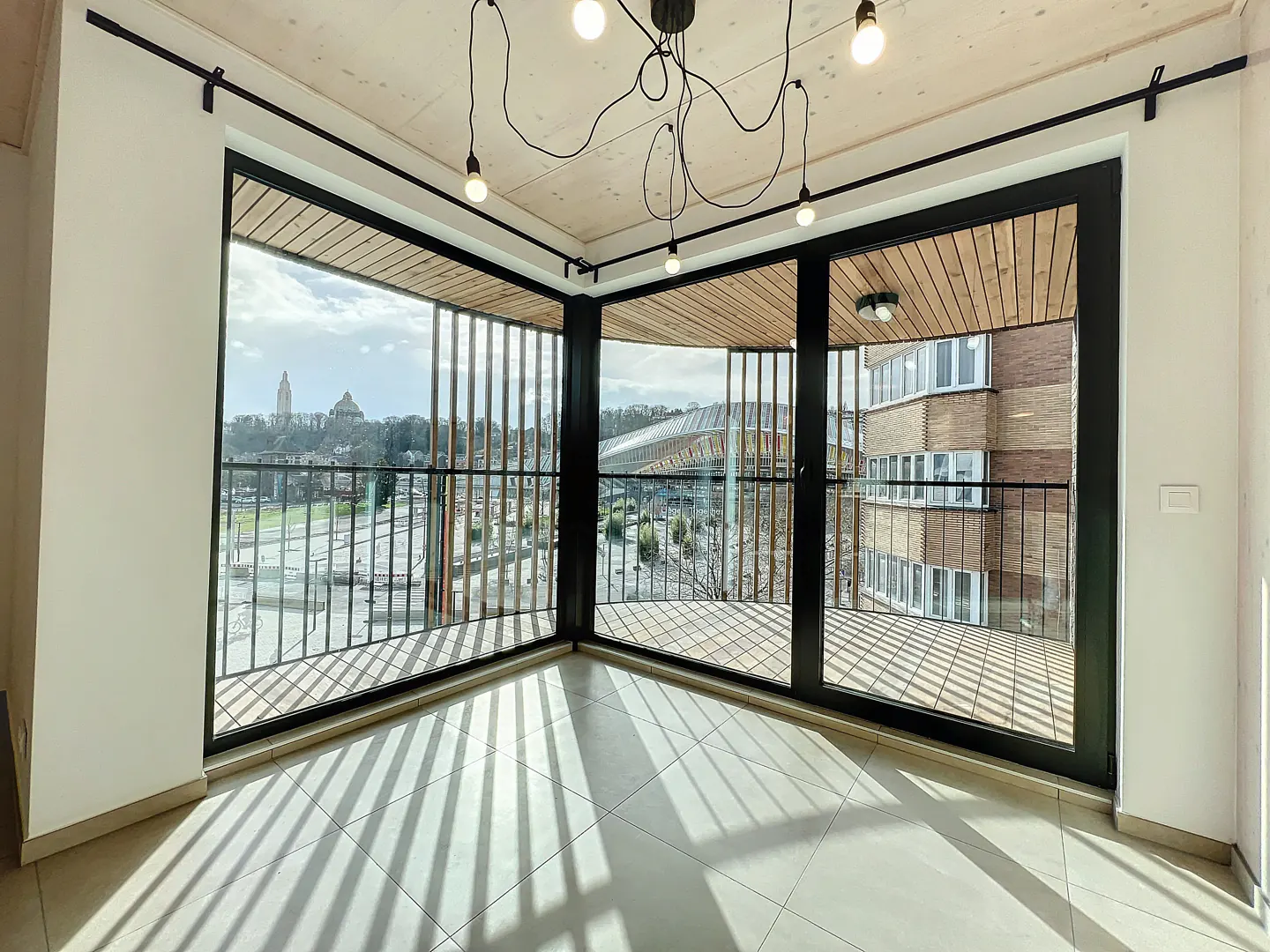 Bright corner room with black-framed windows and a balcony overlooking a city with a cathedral. A modern light fixture hangs from the wooden ceiling.
