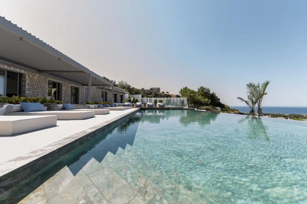 Infinity pool with steps leading into the water, white lounge chairs, stone building, and ocean view under a clear blue sky.