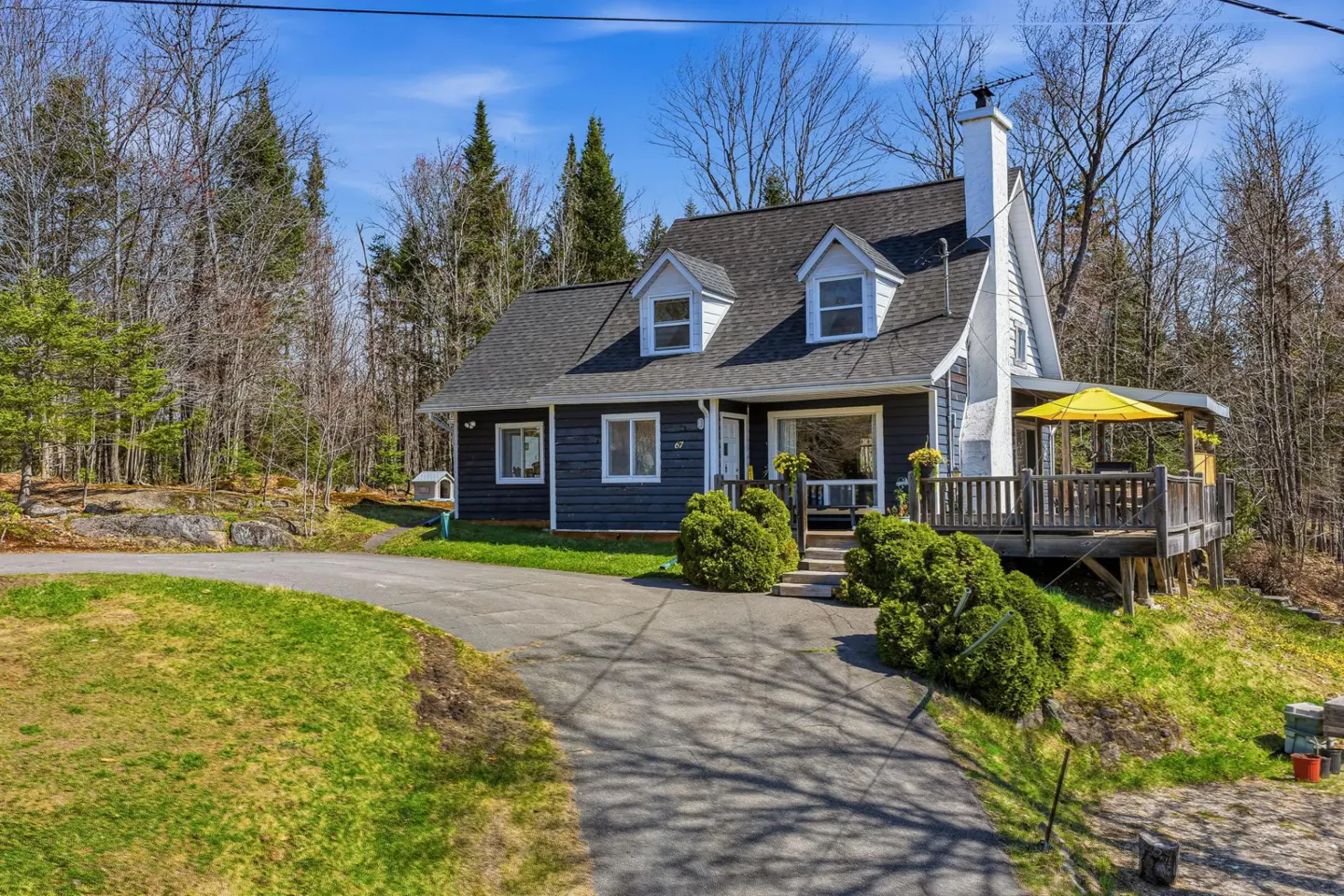 A blue house with a gray roof and white trim sits on a grassy lot with trees in the background. A yellow umbrella is on the deck.