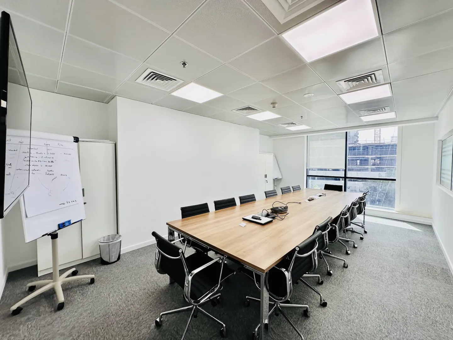 Bright, modern conference room with a long wooden table, black chairs, whiteboard, and large window. The walls are white, and the floor is gray carpet.