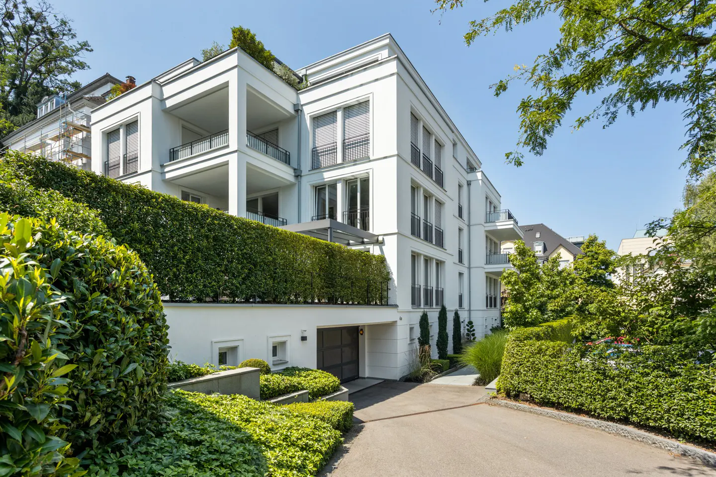 Exterior of a modern white building with balconies, green hedges, and a driveway.