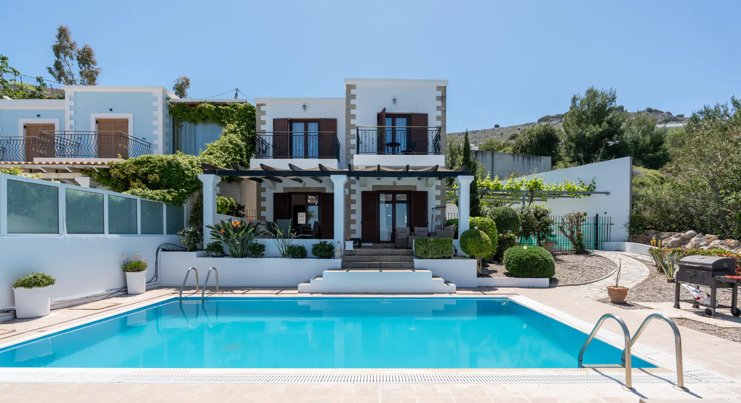 A two-story white house with brown shutters overlooks a turquoise pool on a sunny day. Lush greenery surrounds the property.