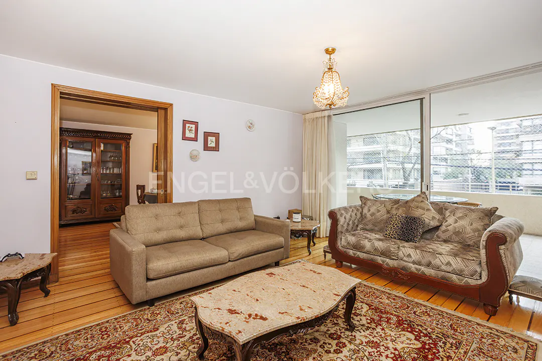 A living room with two sofas, a coffee table, and a view of a balcony. The room has wooden floors and a chandelier.