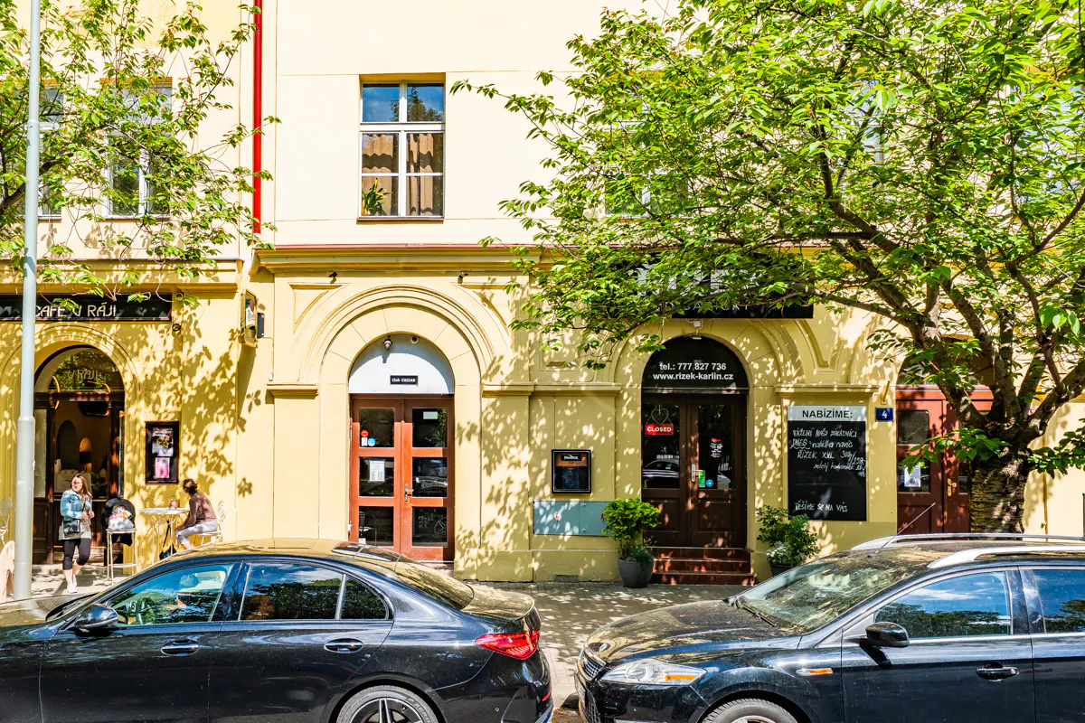 Yellow building facade with arched storefronts, a restaurant entrance, and parked cars on a sunny street.