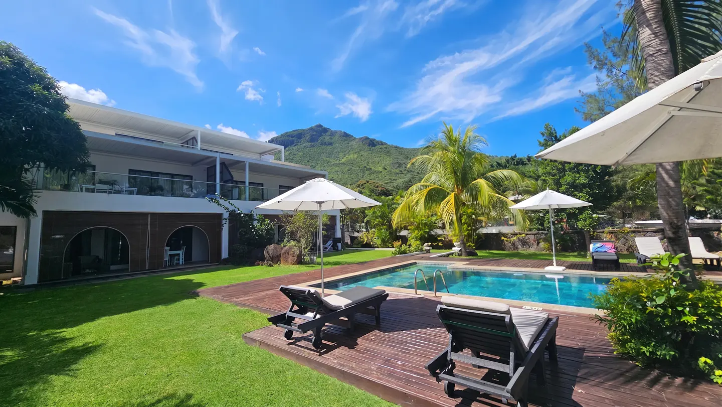 Luxury villa with a pool, lounge chairs, and umbrellas on a wooden deck. Lush green lawn and palm trees surround the pool. Mountain in the background.