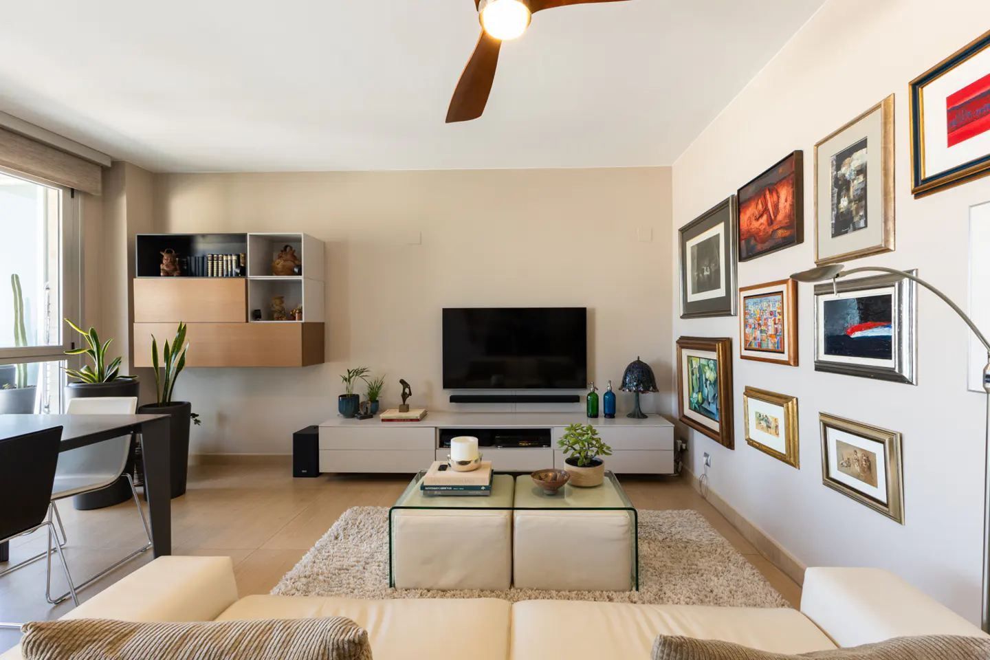 Living room with white sofa, glass coffee table, TV, and gallery wall. A ceiling fan is visible.