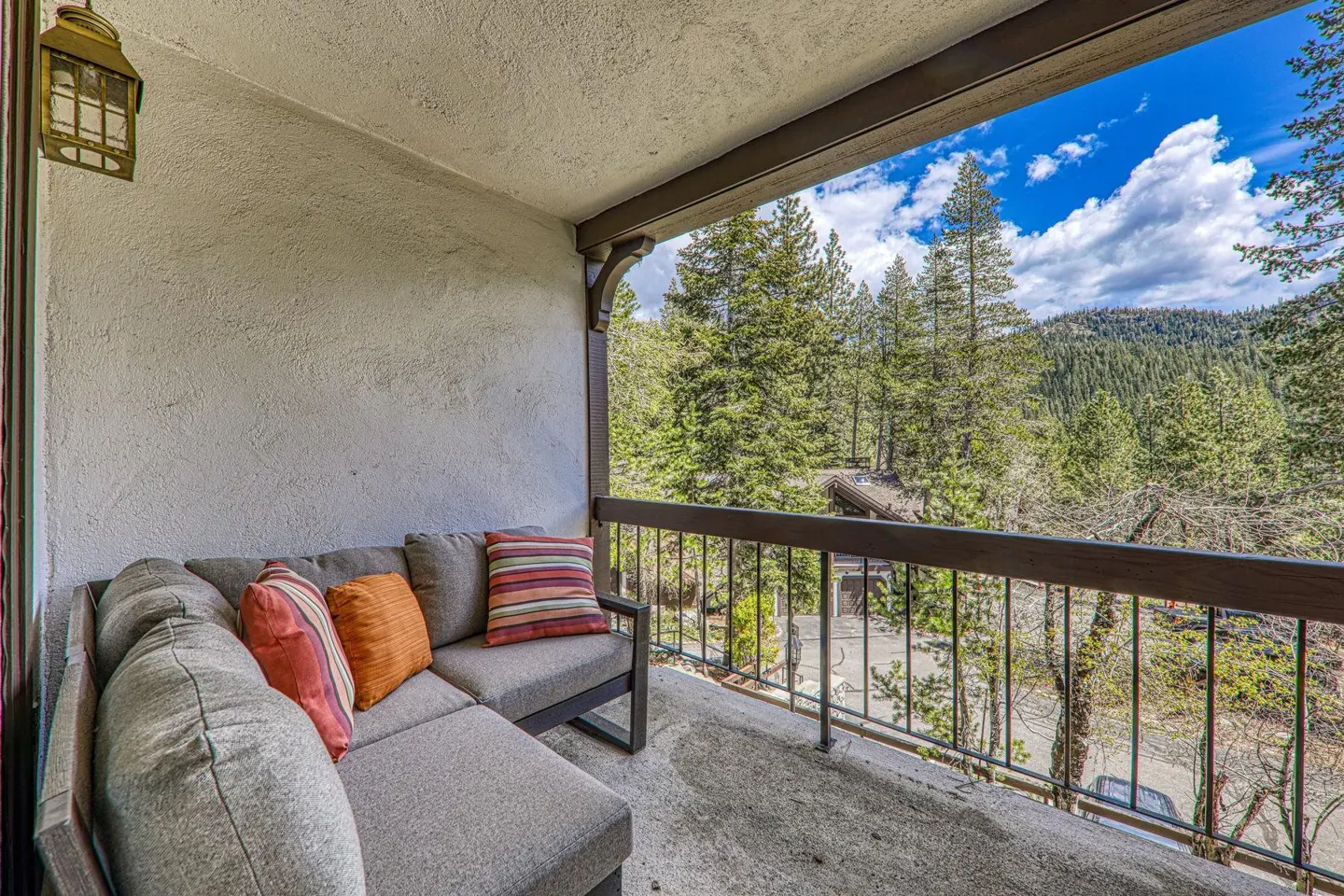 Balcony with gray sectional sofa, colorful pillows, and a view of trees and mountains under a blue sky.