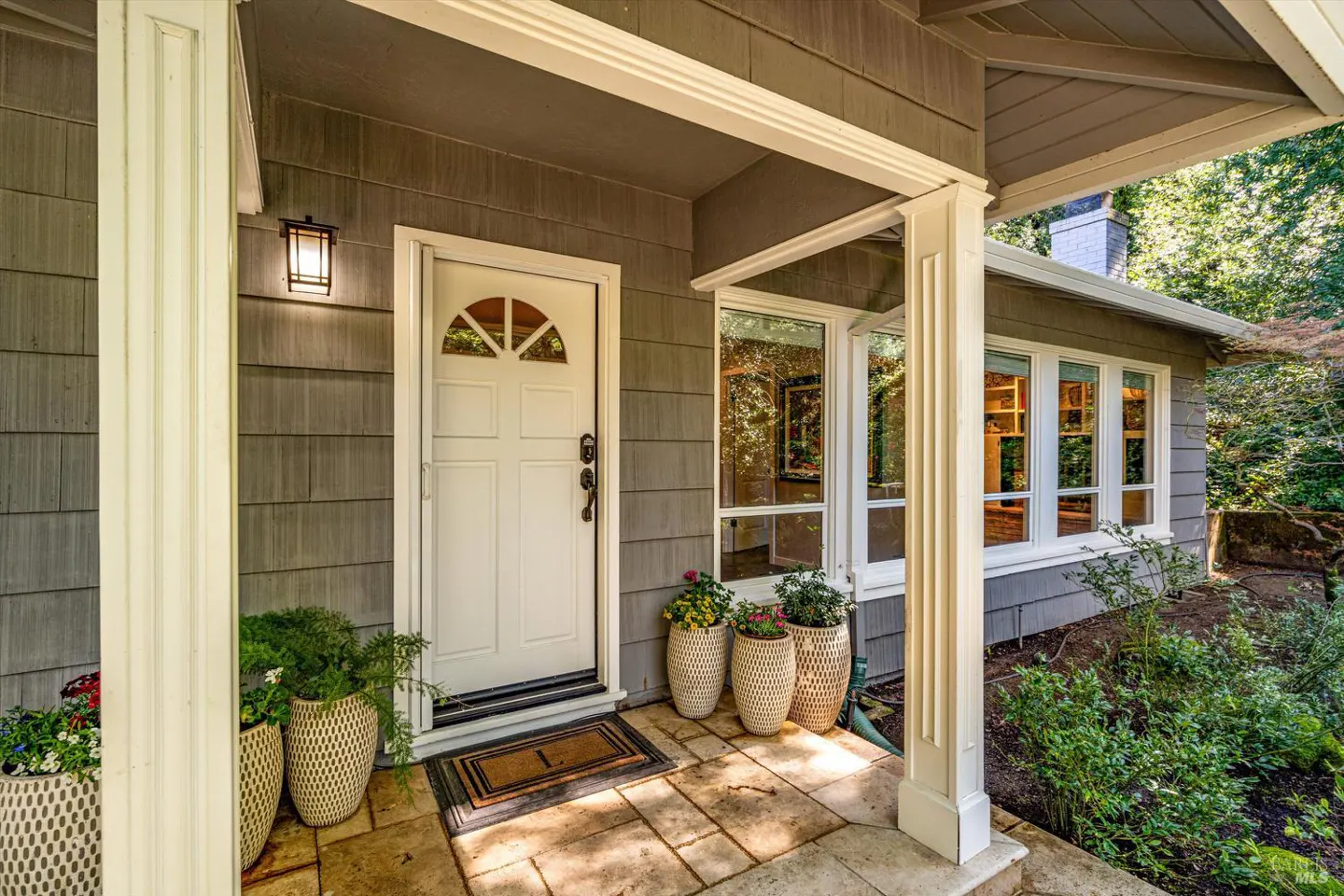Front porch of a gray house with a white door, columns, and potted plants.
