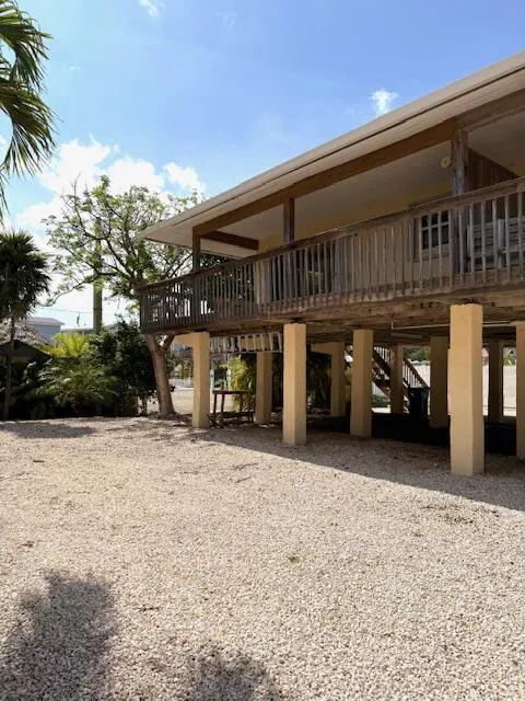 Two-story stilt house with a wooden balcony, beige pillars, and a gravel yard under a blue sky.