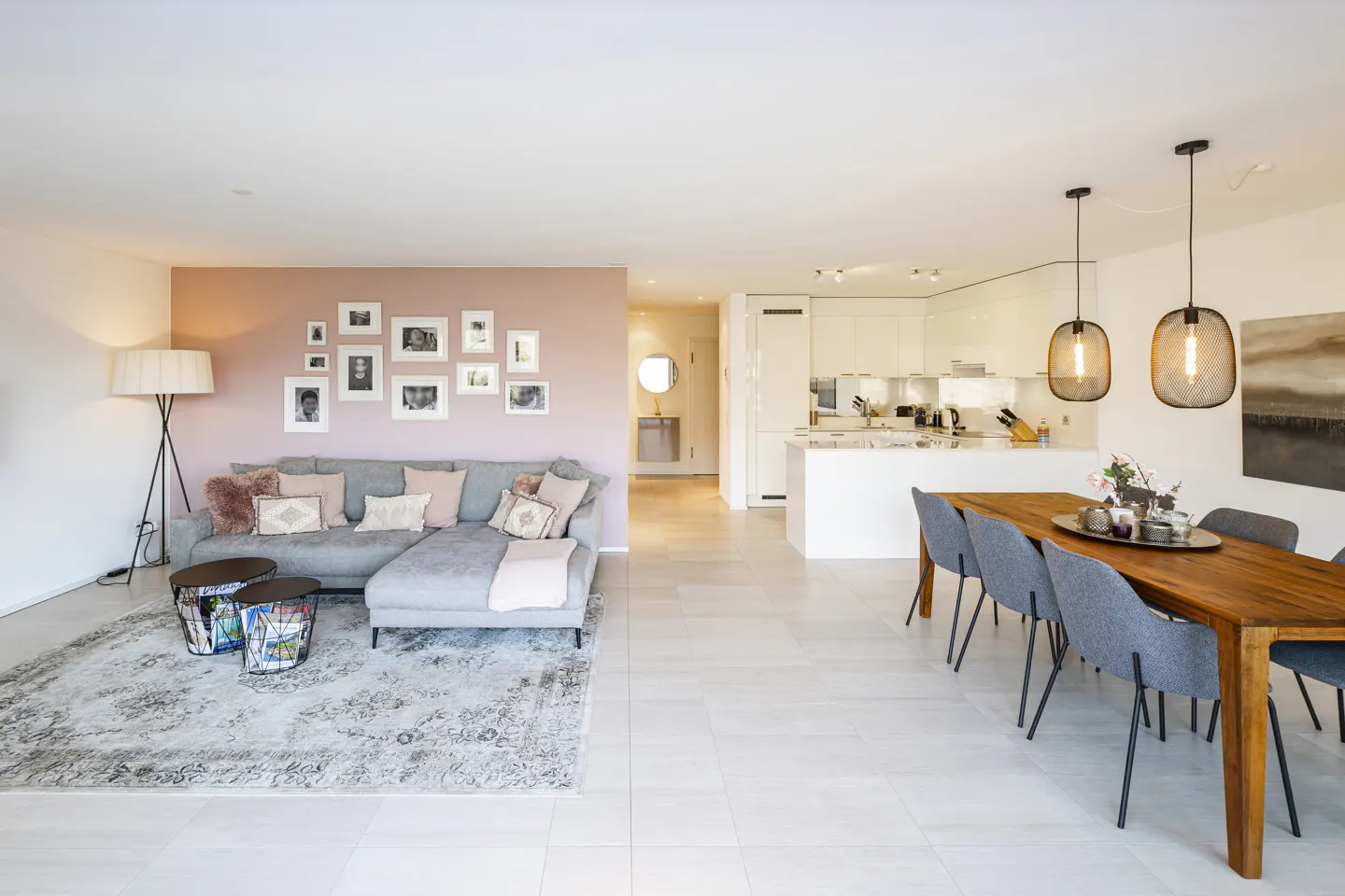 Open-concept living space with a gray sofa, pink accent wall with photos, and a dining table with gray chairs.