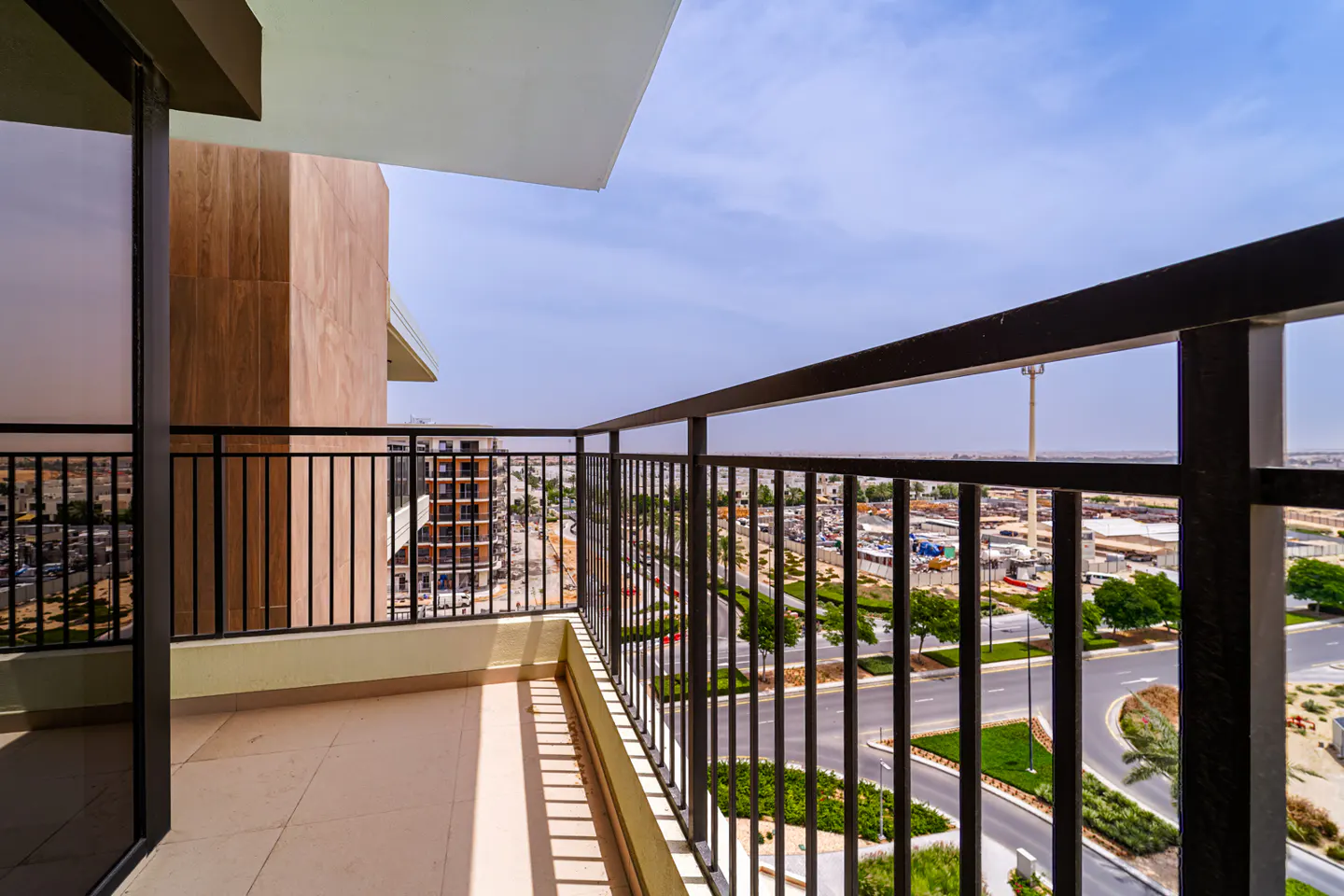 View from a balcony with black metal railings, overlooking a city with roads, buildings, and green spaces under a blue sky.