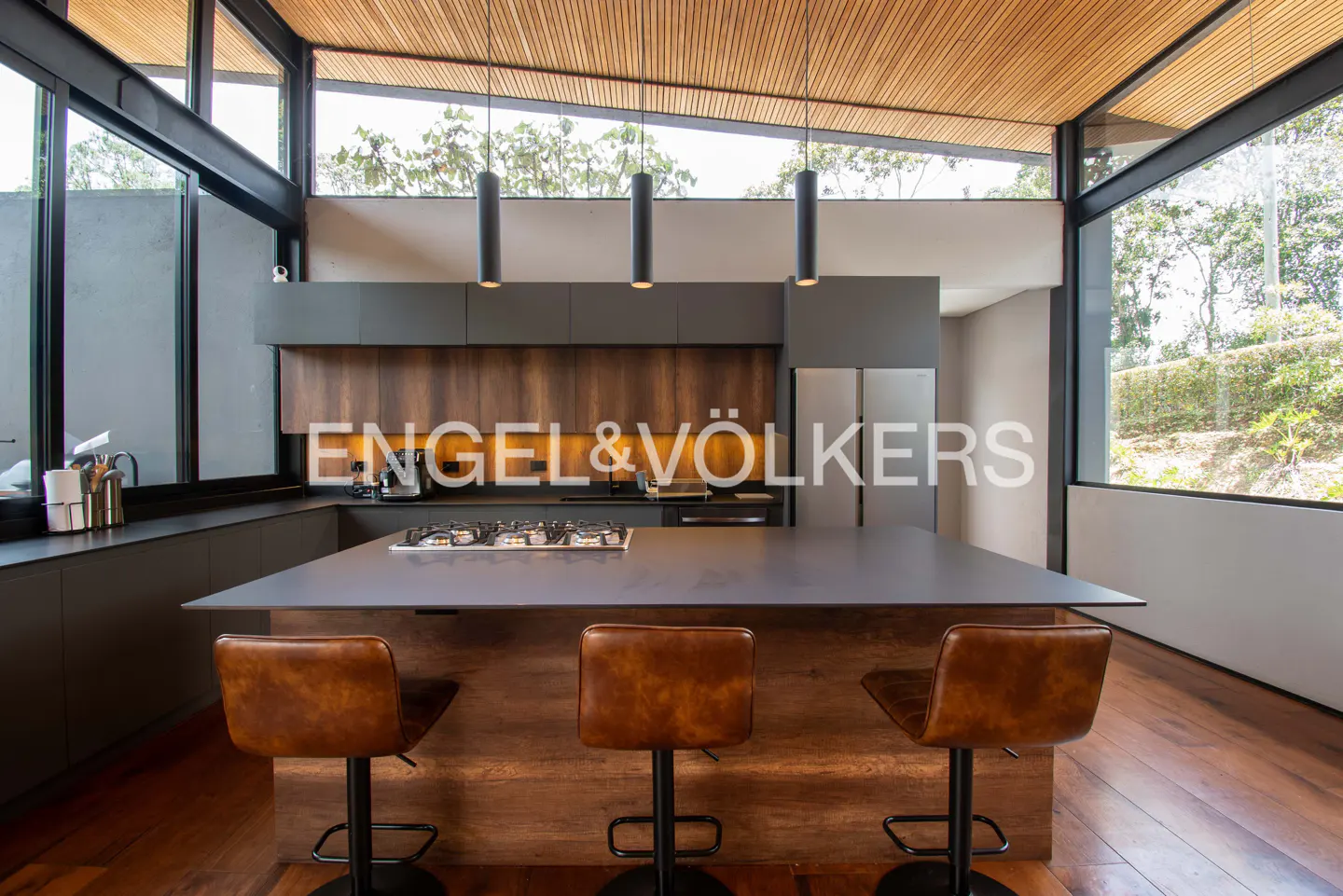 A modern kitchen with wood floors, a gray island with three brown leather stools, and large windows.