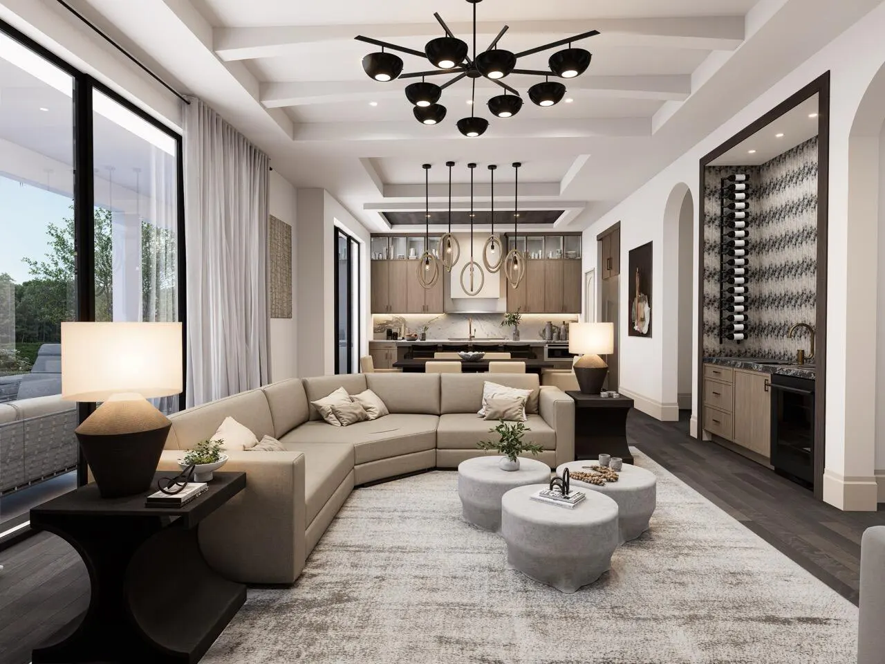 Open-concept living room with a beige sectional sofa, round ottomans, and a black chandelier. A kitchen and wine bar are in the background.