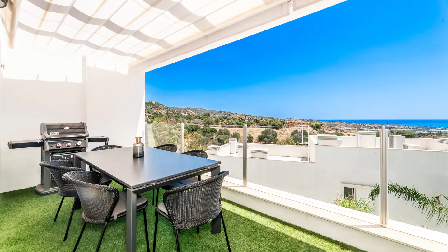 Outdoor patio with a black table, six chairs, and a grill on green turf. Ocean and blue sky in the background.