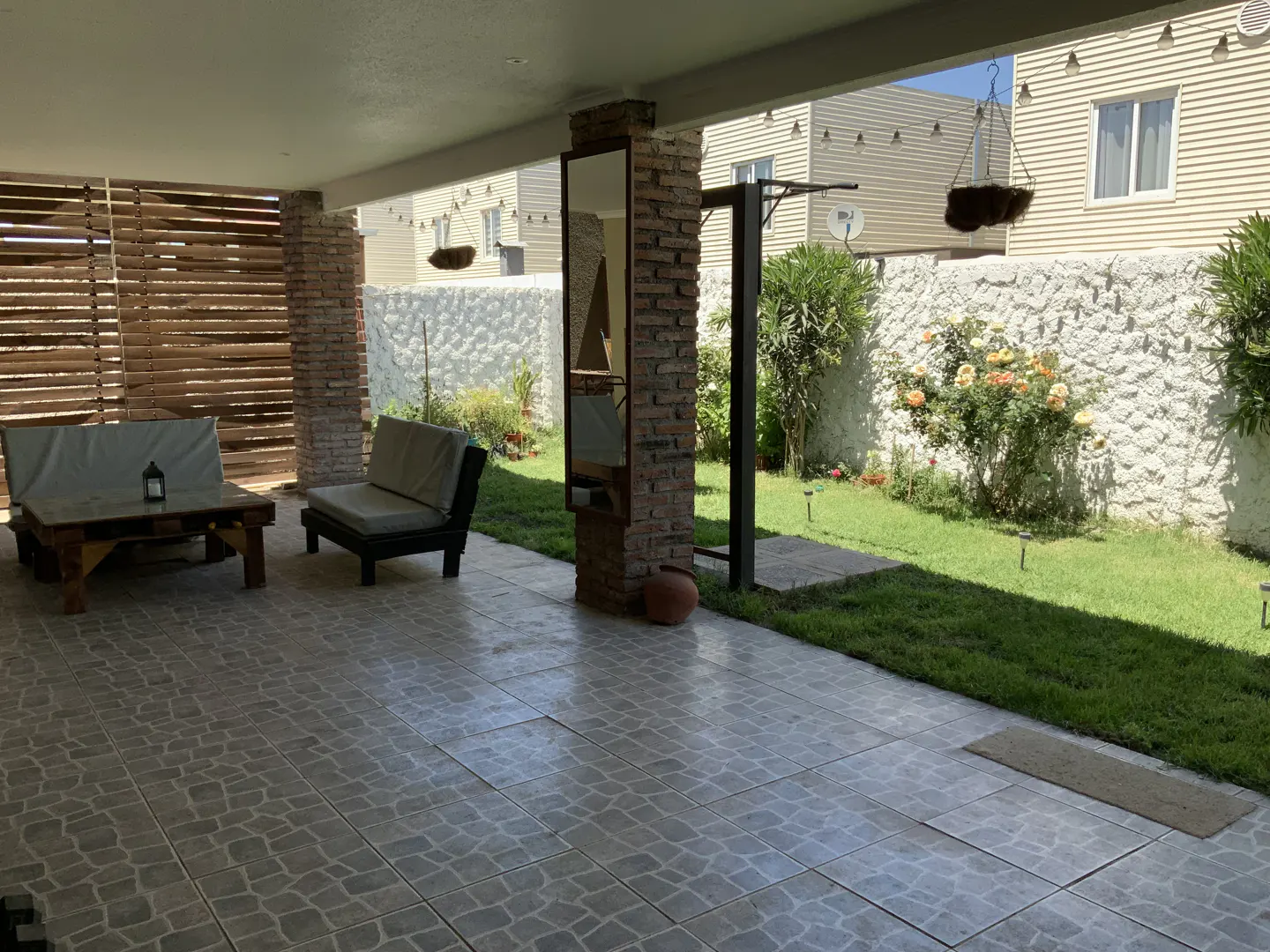 Covered patio with gray tile, wooden furniture, and a brick column with a mirror. A green lawn and white stone wall are visible in the background.