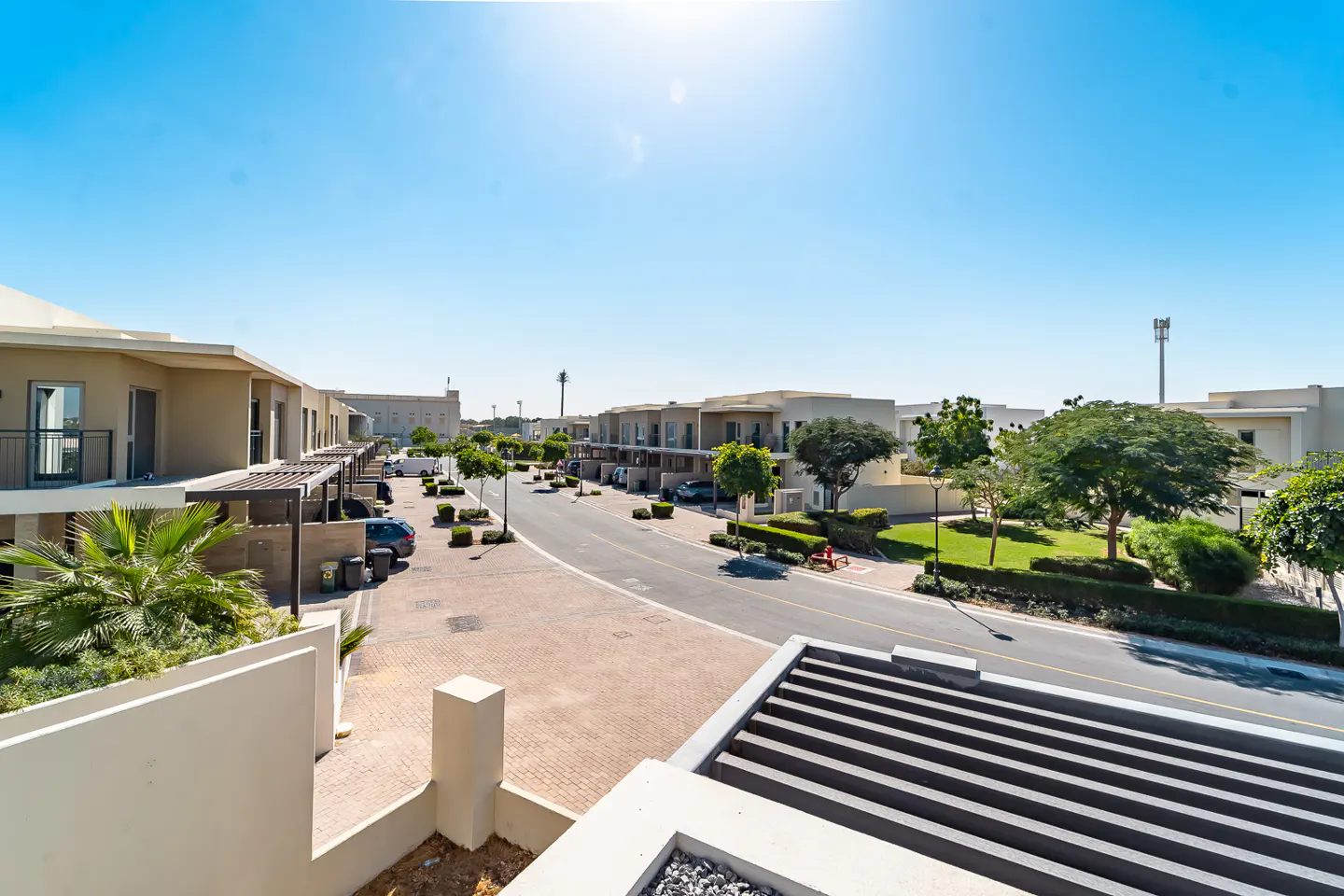 View of a residential street with beige townhouses, green trees, and a clear blue sky. A brick-paved road winds through the neighborhood.