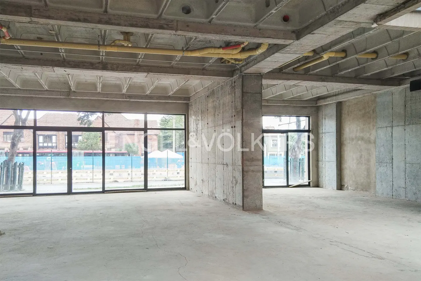 Unfinished commercial space with concrete floors, walls, and ceiling. Large windows offer street views. Yellow pipes run along the ceiling.