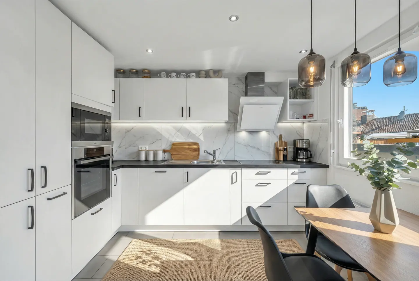 Bright, modern kitchen with white cabinets, black countertops, and marble backsplash. A wooden table with black chairs sits on a woven rug.